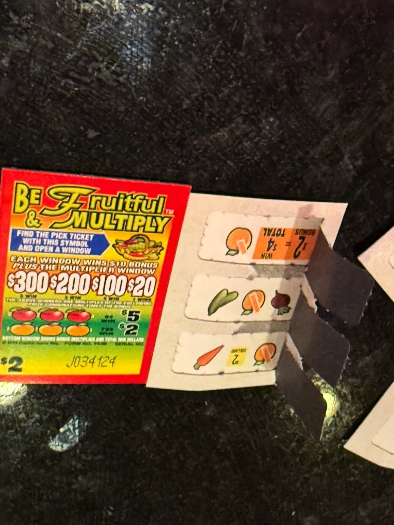 Two pull tabs side by side on a black counter, with one open upside down with a $4 win, the other detailing the name of the pull-tab theme as Be Fruitful and Multiply