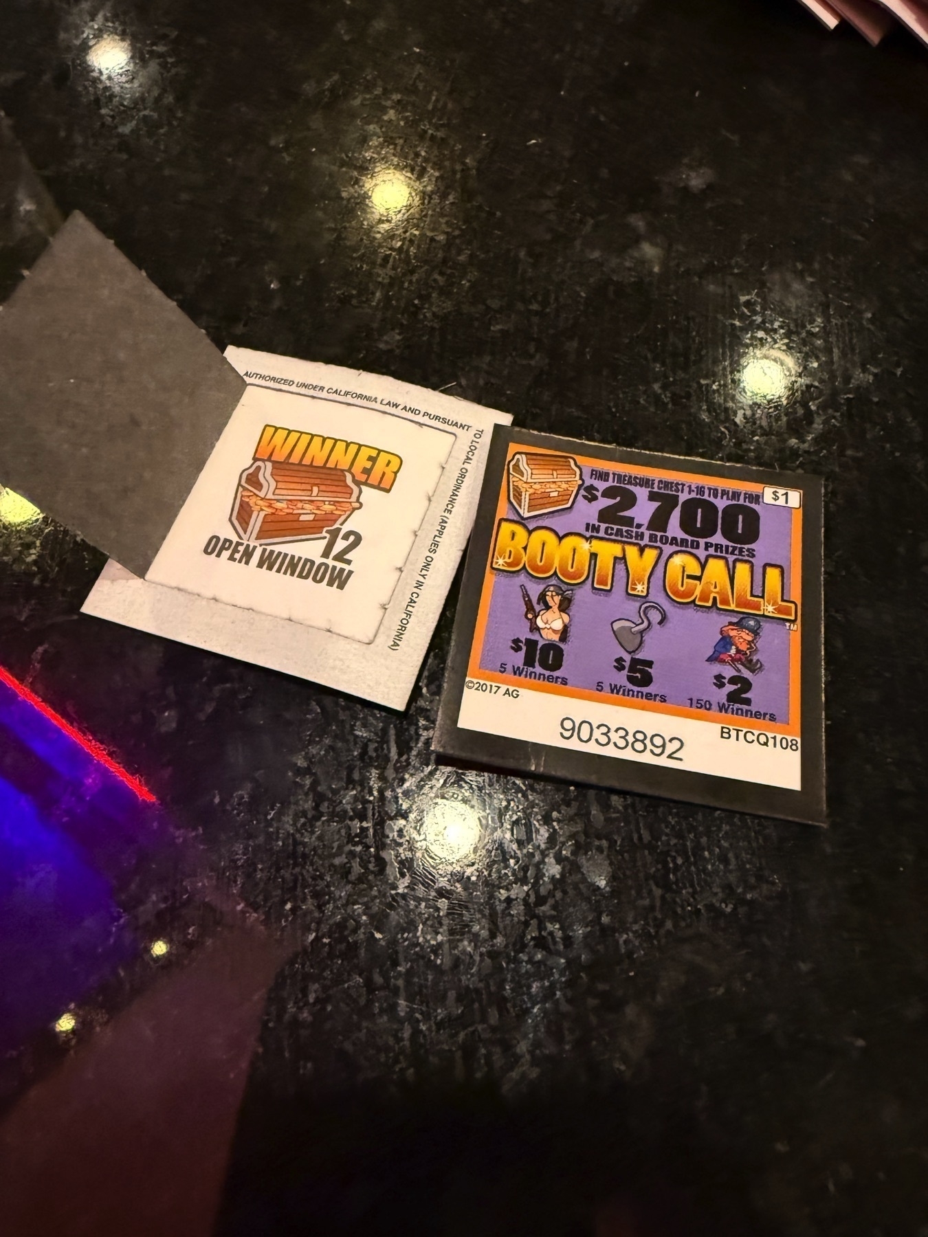 Two single-panel pull tabs side by side on a black counter, with one open as a winner and the other detailing the name of the pull-tab theme as Booty Call
