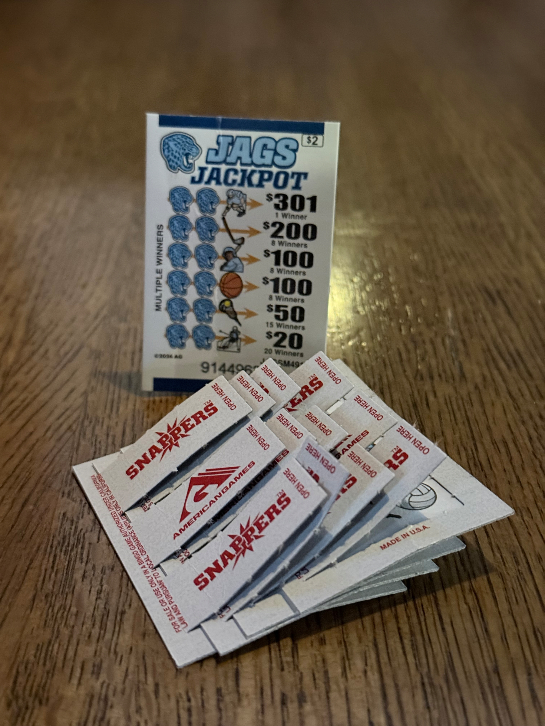 stack of Snappers branded pull-tabs on wooden surface with one upright showing images of a blue-color jaguar logo and various objects like a baseball player, basketball, hockey
