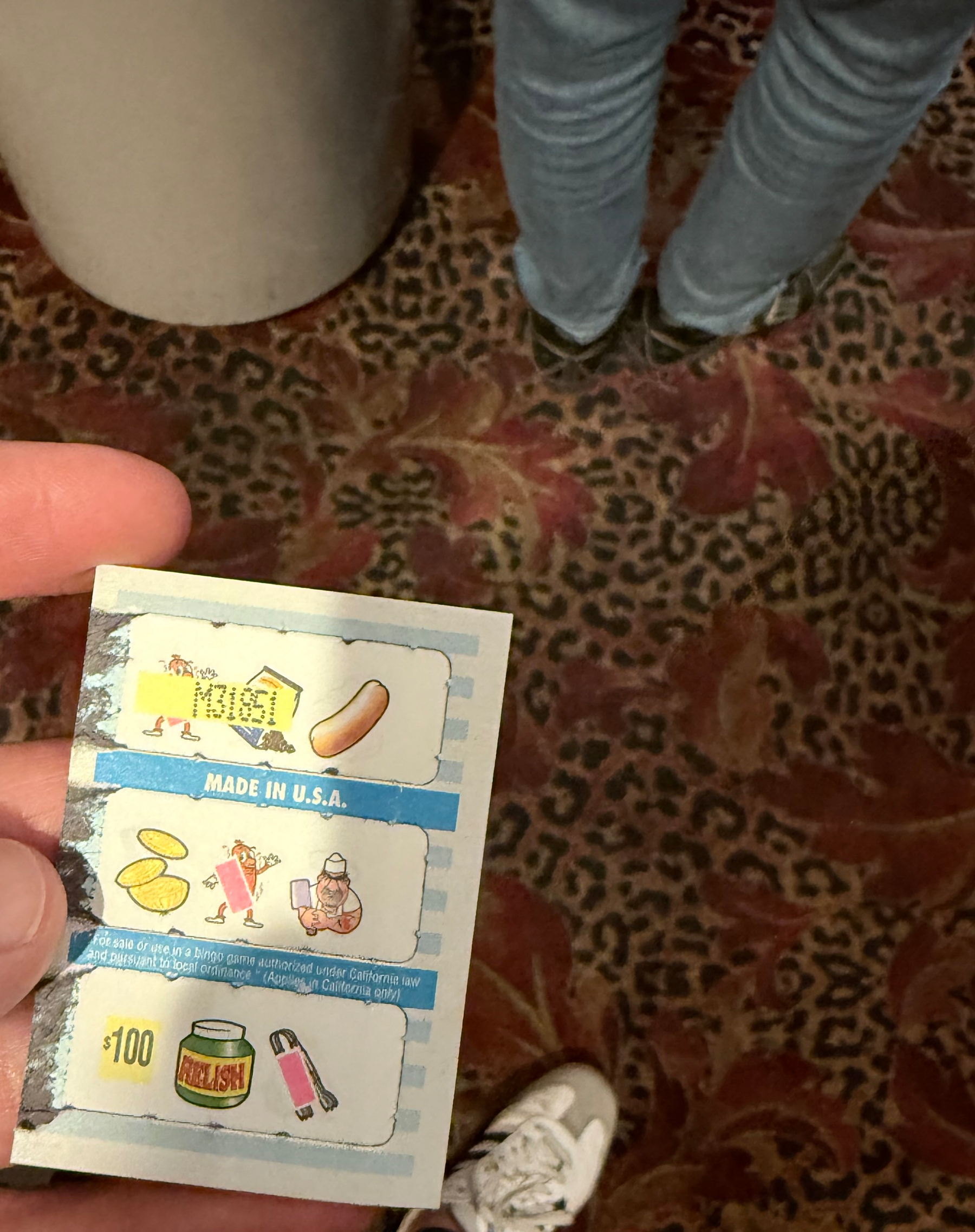 a pull tab held in a hand with $100 winner announced across images of a chef, wiener hot dog, bun, and relish bottle